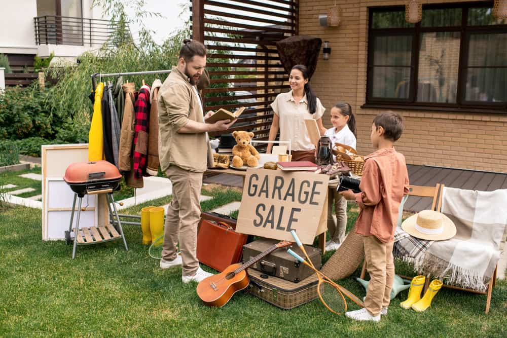 Man checking out a book at a yard sale. Woman with kids wait for customers.